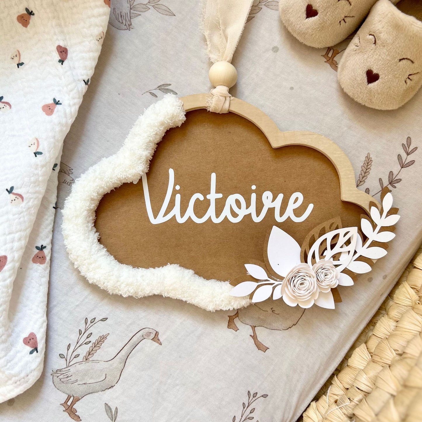 Décoration murale bébé fille personnalisée avec le prénom de l'enfant, nuage aux tons marron et écru, déposé dans un couffin avec un lange et des petits chaussons 1 mois.
