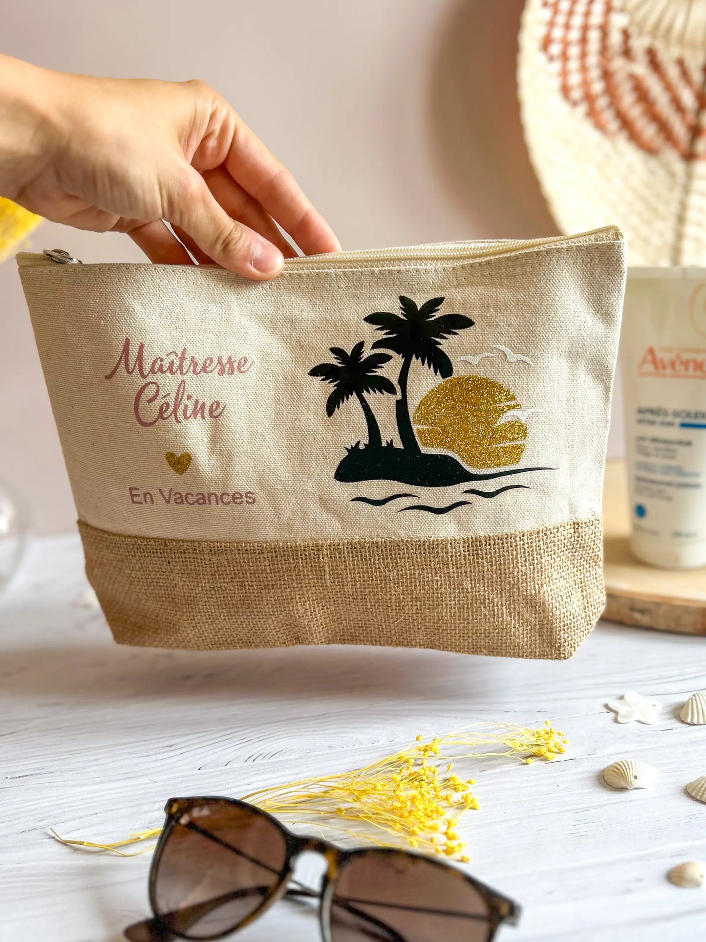 Trousse personnalisée en coton et toile de jute avec l'inscription "Maîtresse Céline en vacances", posée sur une table avec lunettes, crème solaire et coquillages.