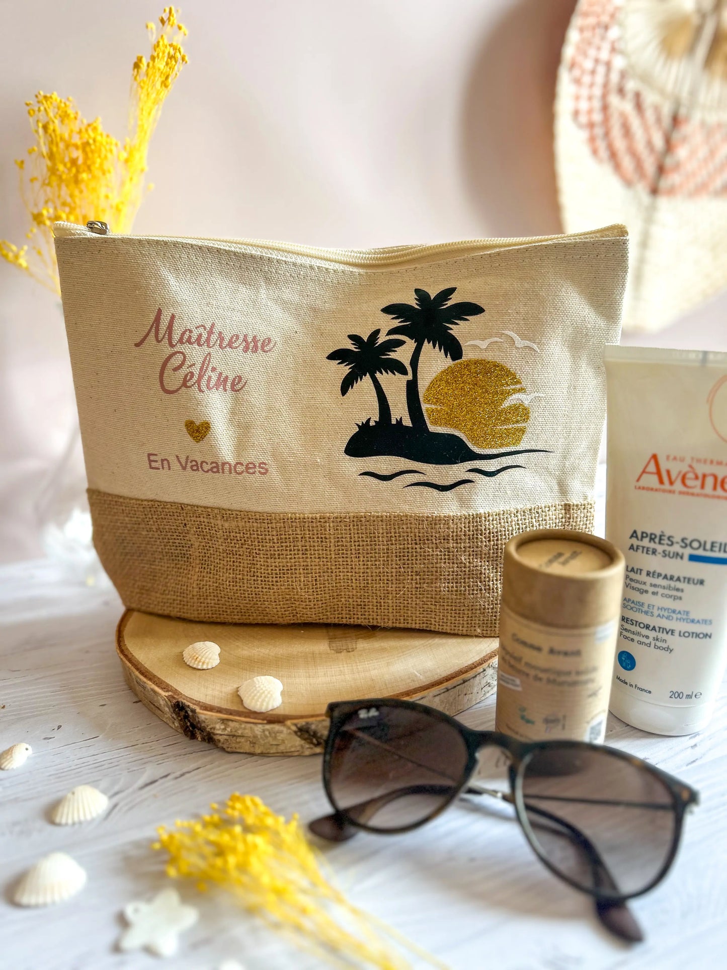 Trousse en coton et jute avec motif palmier et texte "Maîtresse Céline en vacances", mise en scène estivale avec fleurs jaunes et lunettes.