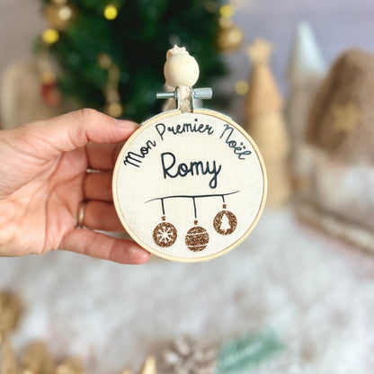 Boule de  Noël fait main et personnalisée au prénom de bébé, tenue à la main devant un décor de Noël.