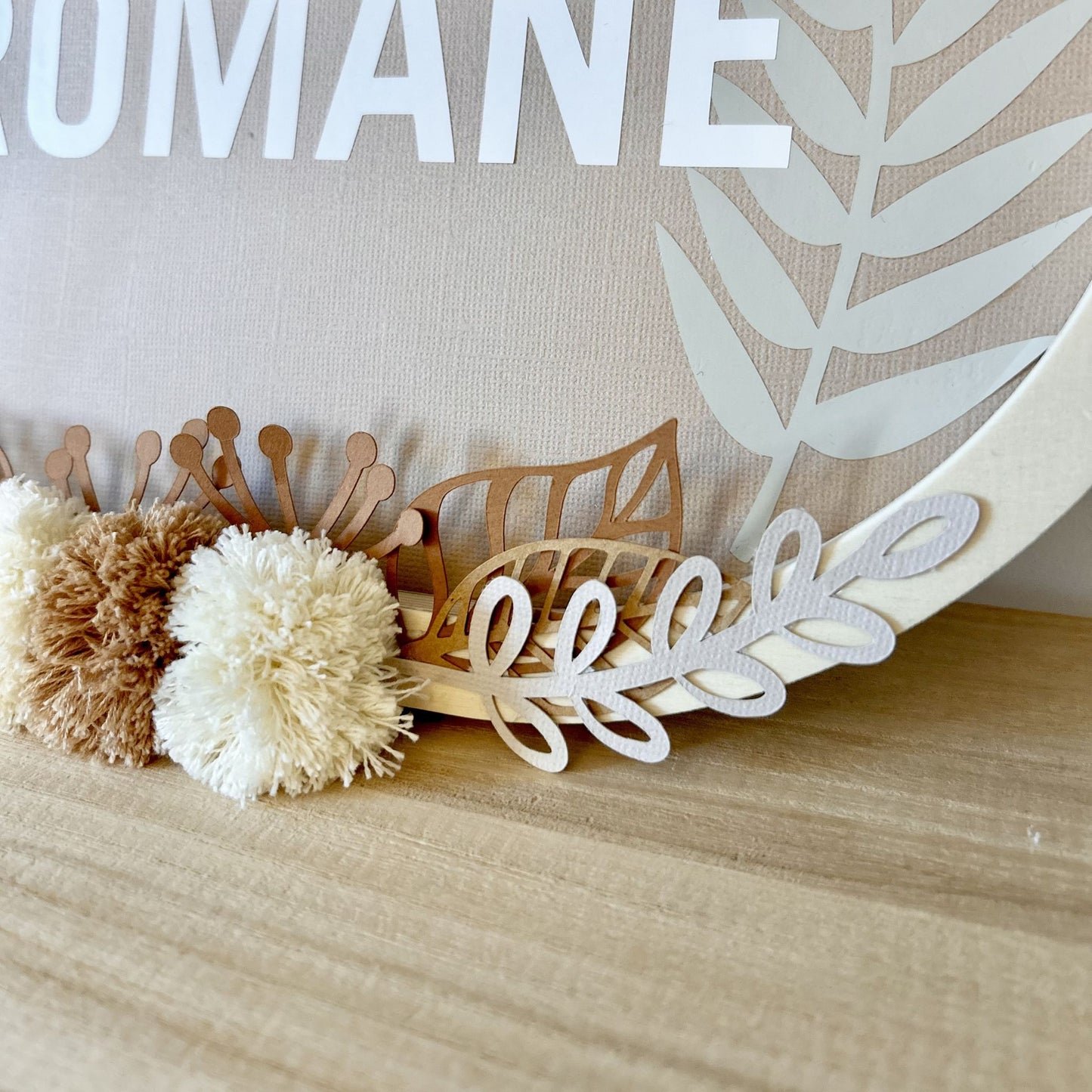 Détail des pompons en laine Oeko-Tex et du feuillage en papier sur cadre ovale personnalisé avec le prénom.