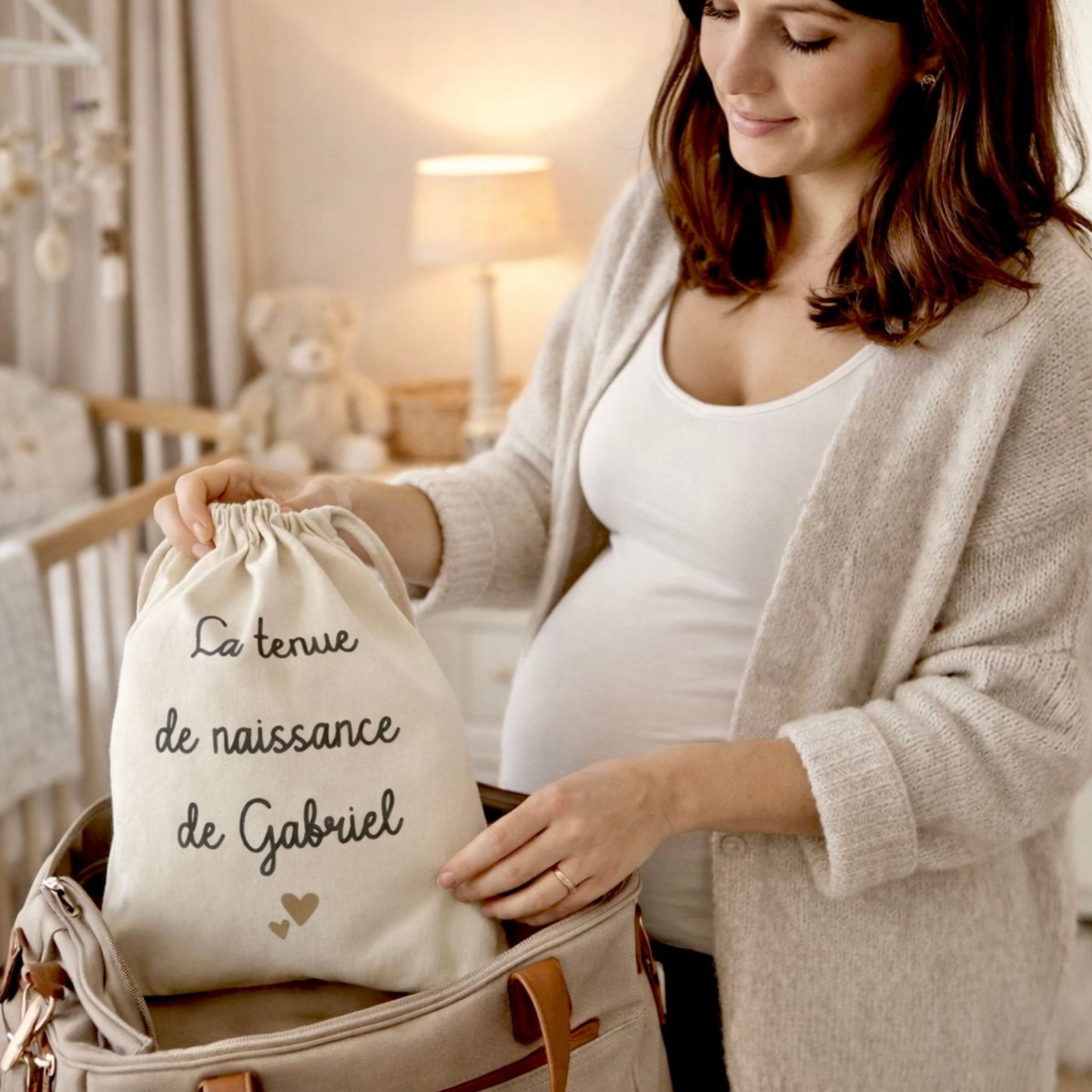 Femme enceinte en train de préparer sa valise de maternité et d’y glisser un pochon en coton contenant la tenue de naissance de bébé.