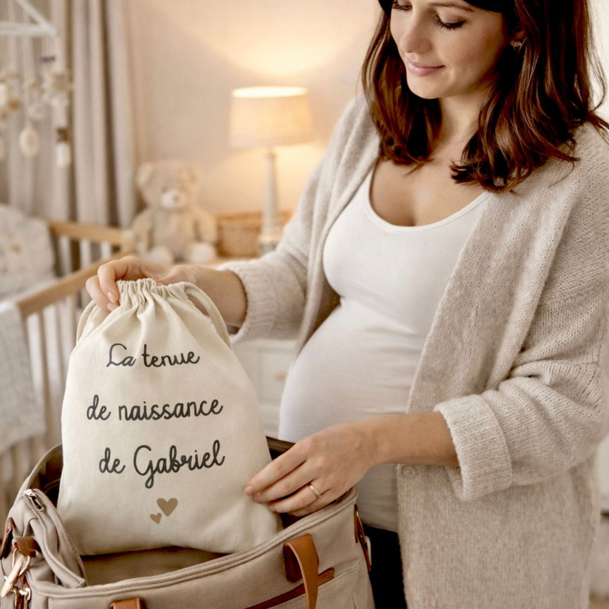 Femme enceinte en train de préparer sa valise de maternité et d’y glisser un pochon en coton contenant la tenue de naissance de bébé.